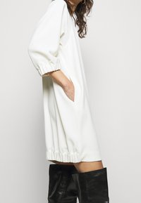 Vit pulloverklänning med trekvartslånga ärmar, sidofickor och rynkad fåll. Tyget verkar vara slätt med avslappnad passform.