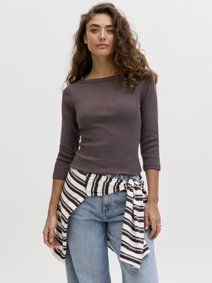 Jeune femme aux cheveux bruns bouclés portant un haut marron à manches trois-quarts, un jean bleu clair et une chemise rayée nouée autour de la taille.