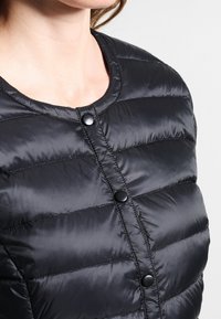 Černá péřová bunda s kulatým výstřihem, horizontálním prošíváním a černými patentními knoflíky vpředu. Hladká, lesklá textura látky.