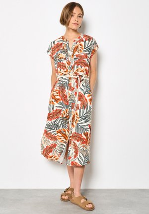 Młoda kobieta stojąca na tle białej ściany, ubraną w midi sukienkę na guziki z pomarańczowo-zielonym wzorem liści oraz beżowe sandały na otwarty palec.