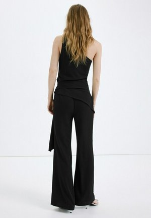 Mujer con cabello largo y ondulado, vestida con un top negro de un solo hombro y pantalones negros de pierna ancha, de pie frente a un fondo blanco liso.