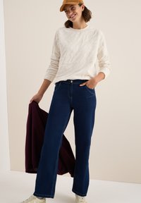 Pull en texture crème, jeans ample bleu foncé avec poches avant et baskets blanches. Tenant une veste bordeaux, debout contre un fond clair.