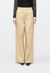 Beige vida byxor med slät textur, resår i midjan och två sidofickor, kombinerade med svarta spetsiga klackar.