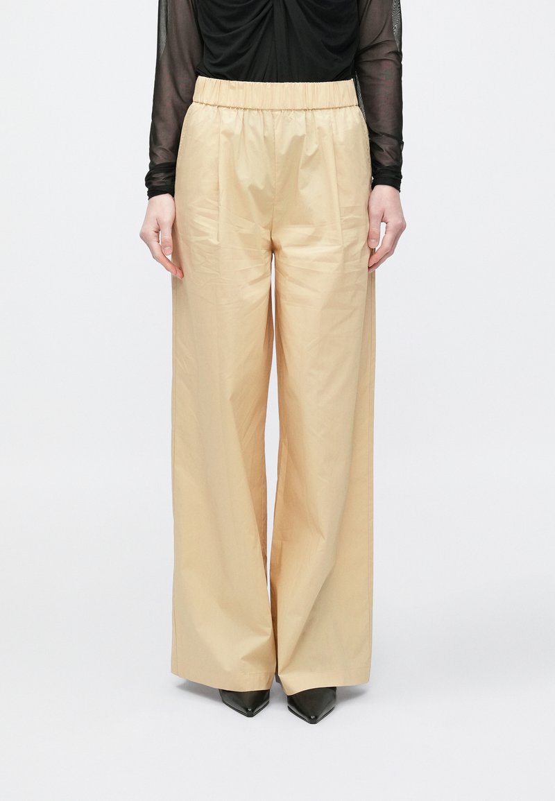 Beige vida byxor med slät textur, resår i midjan och två sidofickor, kombinerade med svarta spetsiga klackar.