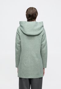 Lysegrønn kåpe med teksturert finish, lange ermer og stor hette. Designet har en rett passform og sømmer bak for struktur.