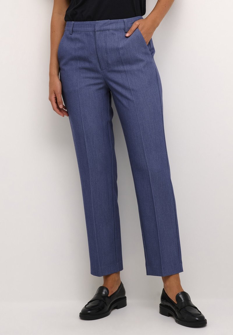 Femme portant un pantalon bleu droit avec des plis marqués, des mocassins noirs et un haut noir, debout sur un fond blanc.