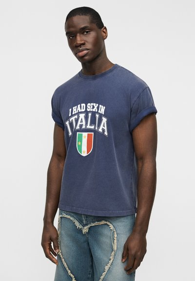 Μπλούζα από βαμβάκι σε ναυτικό μπλε με σχεδιασμό με ρεβέρ στα μανίκια και τυπωμένο κείμενο με γραφικό ενός ασπίδας σε πράσινο, λευκό και κόκκινο.