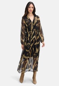 Eine Frau trägt ein schwarzes, durchsichtiges Midikleid mit goldenen botanischen Mustern und langen Ärmeln, kombiniert mit hellbraunen Wildlederstiefeln.