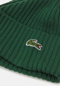 Green ribbed knit beanie hat made of wool with a folded cuff and a small embroidered crocodile logo in green and red.