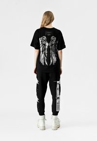 Camiseta negra con gráficos de alas plateadas en la parte trasera, combinada con joggers negros que exhiben un diseño impreso y acentos en blanco, llevada con botas blancas.