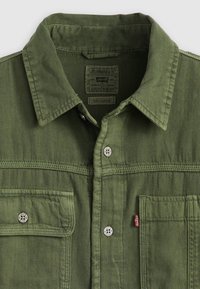 Chemise en denim vert olive avec un col pointu, des boutons-pression en argent et deux poches sur la poitrine avec des accents de marque. Tissu texturé.