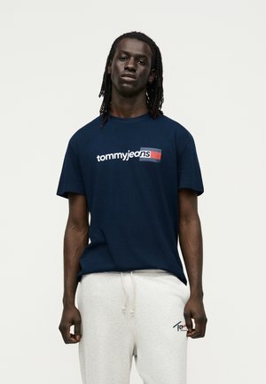 Mand med dreadlocks iført en mørkeblå "tommy jeans" t-shirt og lysegrå joggingbukser, der står mod en ensfarvet baggrund.