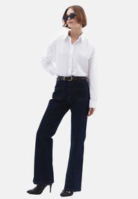 Witte blouse met knopen, donkerblauwe flared jeans met zichtbare naden, bruine leren riem en zwarte hoge hakken.