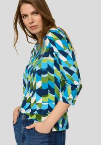 Frau trägt ein farbenfrohes Oberteil mit geometrischem Muster aus blauen, grünen und weißen Formen, kombiniert mit blauen Jeans, Hände in den Taschen.
