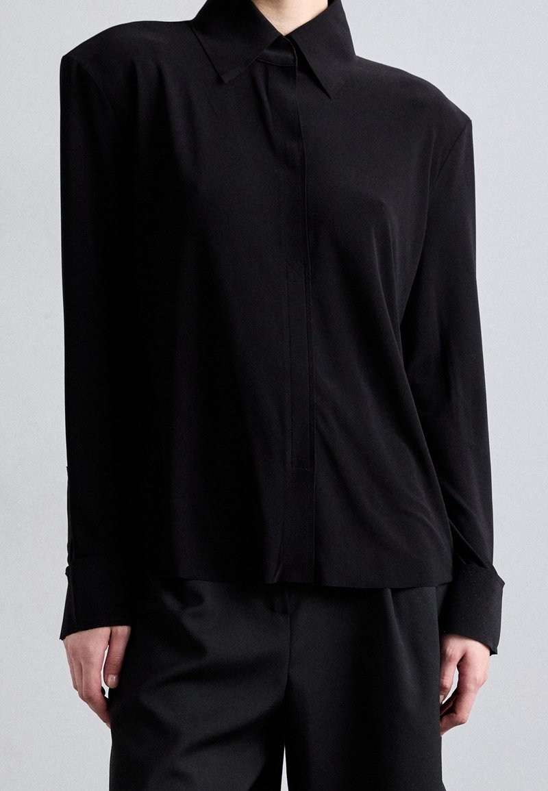 Black long-sleeve shirt with a pointed collar, front button closure, and wide cuffs. Smooth fabric with a relaxed fit.