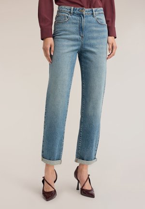 Frau trägt hellblaue Straight-Leg-Jeans mit Umschlag, kombiniert mit einem bordeauxfarbenen Langarmshirt und passenden hochhackigen Schuhen mit gekreuzten Riemen.