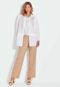 Camicia bianca con dettagli dorati intricati, abbinata a pantaloni beige a gamba larga e sandali con piattaforma di colore chiaro.