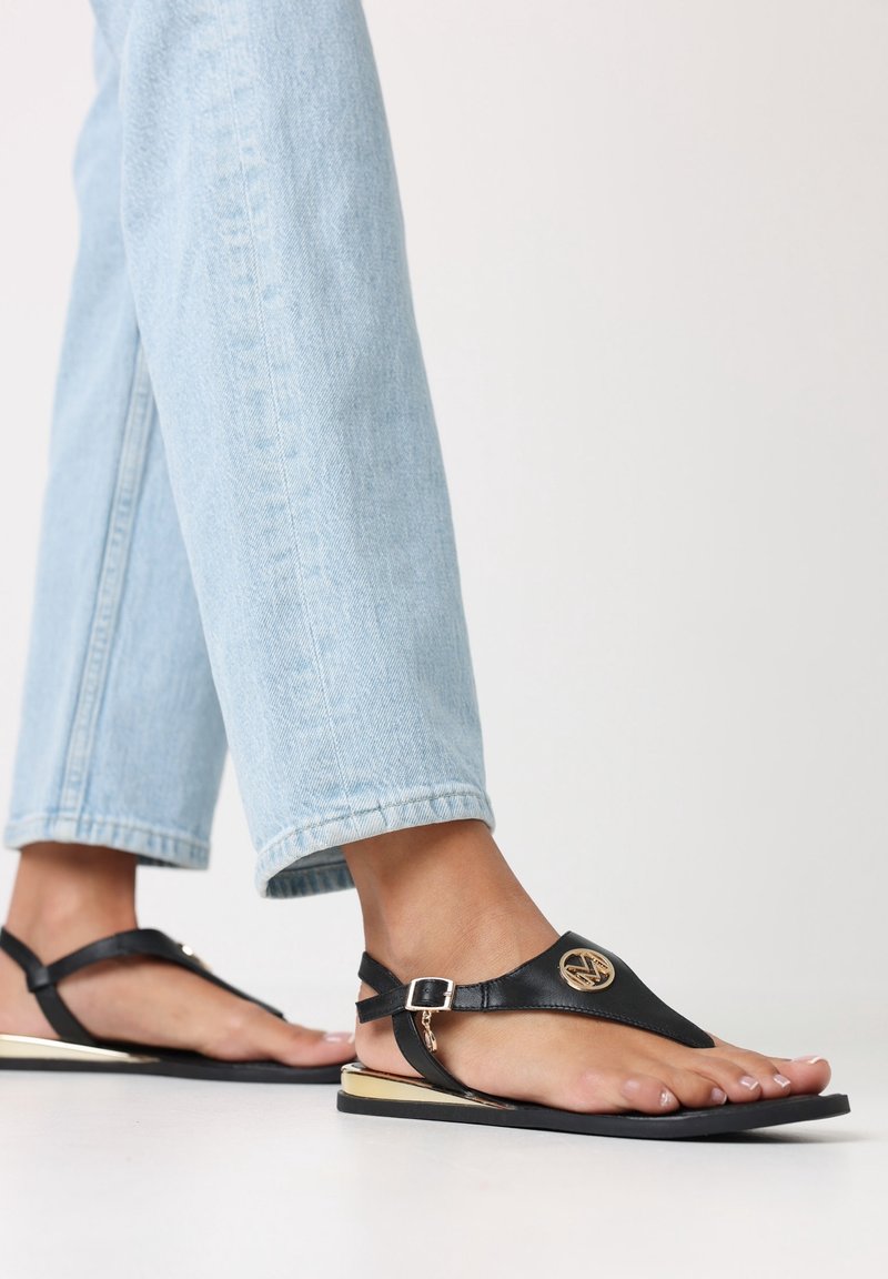 Sandales en cuir noir avec un design à bride en T et un accent circulaire doré, dotées d'une semelle plate et d'une boucle à la cheville. Portées avec un jean clair.