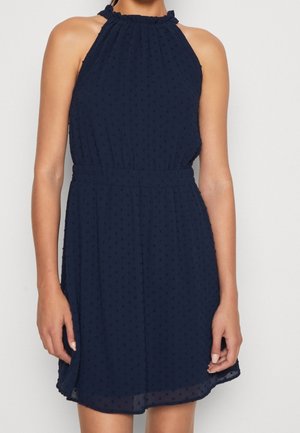 Vrouw draagt een mouwloze marineblauwe halterjurk met een gestructureerd patroon van kleine stippen en een elastische tailleband, staand tegen een effen achtergrond.