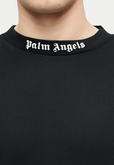 Prim-plan cu o persoană purtând un tricou negru cu guler rotund, pe care este inscripționat "Palm Angels" cu font gotic alb.