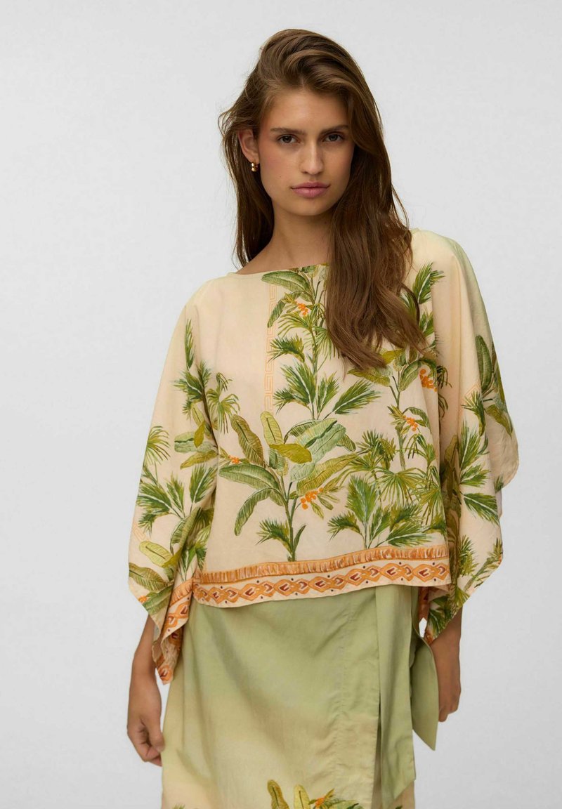 Femme aux longs cheveux bruns portant un haut beige ample à motif de feuilles et une jupe vert clair sur un fond gris uni.
