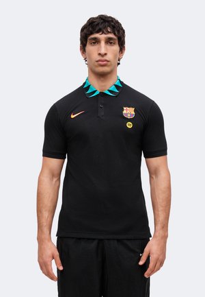 Άνδρας με μαύρο πόλο μπλουζάκι FC Barcelona με γιακά σε τιρκουάζ και μαύρο, πορτοκαλί λογότυπο Nike και κίτρινο λογότυπο Spotify, στέκεται μπροστά σε απλό φόντο.