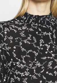 Blouse en tissu noir avec un col froncé, ornée d'un motif floral blanc et de délicates motifs de vigne sur toute sa surface.