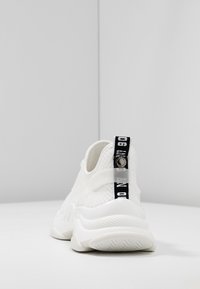 Sneaker bianco con tomaia in rete, suola in gomma e una linguetta nera con marchio sul tallone. Presenta un design testurizzato e uno stile minimalista.