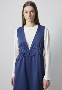Rochie fără mâneci, de culoare albastru marin, cu decolteu adânc în formă de V, talie strânsă și accente contrastante de culoare crem cu șnur. Materialul pare neted și ușor.