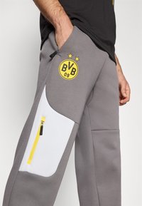 Grå joggebukser har en hvid lomme med en gul lynlås, samt et sort og gult "BVB 09"-logo med to stjerner over det.