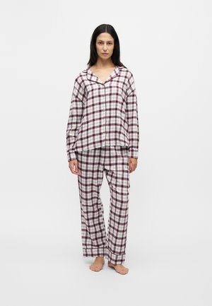Femme debout pieds nus portant un haut de pyjama à carreaux blanc et bordeaux assorti à un pantalon, sur fond uni.