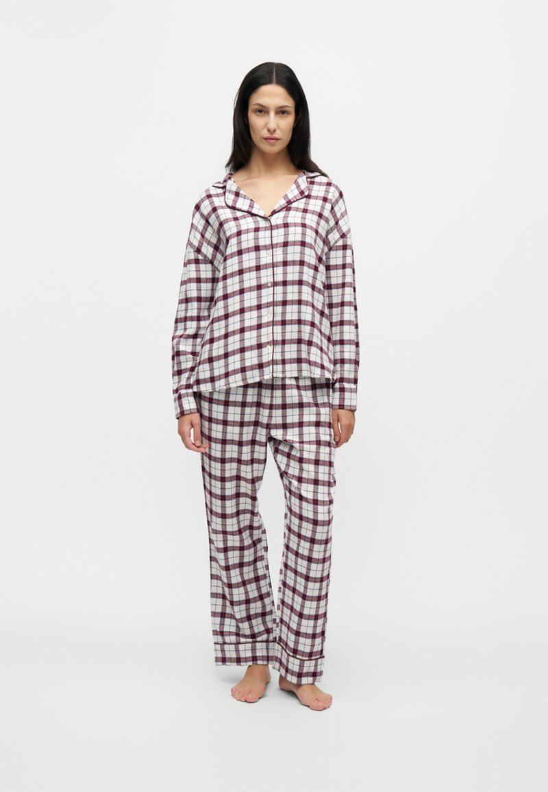 Femme debout pieds nus portant un haut de pyjama à carreaux blanc et bordeaux assorti à un pantalon, sur fond uni.