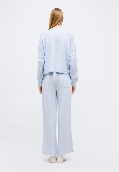 Γυναίκα με μακριά ξανθά μαλλιά που στέκεται, φοράει ανοιχτό μπλε φαρδιά μπλούζα με μακριά μανίκια και φαρδιά παντελόνια, σε οπτική από πίσω.