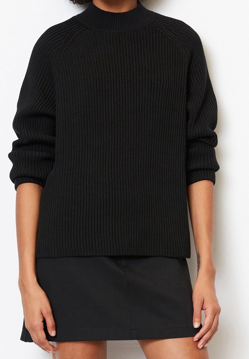 Personne portant un pull en tricot côtelé noir avec les manches retroussées et une jupe noire, debout devant un fond blanc uni.