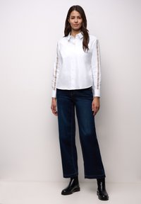 Kvinna som står mot en ljus vägg, iklädd en vit skjorta med knappar och randiga ärmar, mörka vida jeans och svarta ankelstövlar.