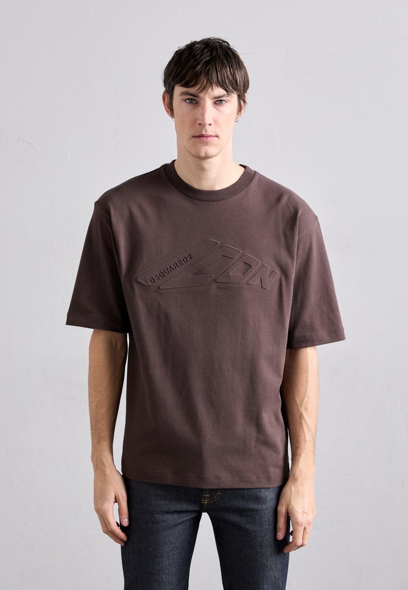 T-shirt de algodón marrón con mangas cortas y cuello redondo, que presenta un logo en relieve en la parte frontal. Combinado con jeans oscuros.