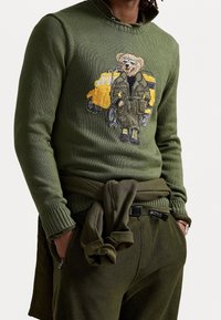 Maglione verde lavorato a maglia con colletto a costine, caratterizzato da un design ricamato di un orso e un veicolo giallo. I dettagli includono una vita e polsini coordinati con fiocco.