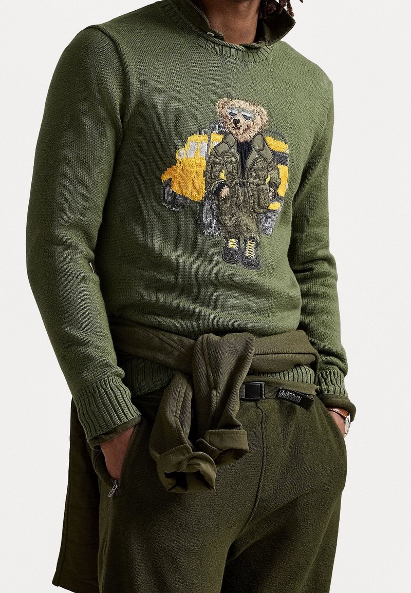 Maglione verde lavorato a maglia con colletto a costine, caratterizzato da un design ricamato di un orso e un veicolo giallo. I dettagli includono una vita e polsini coordinati con fiocco.