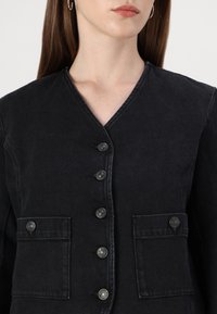 Veste en denim noire avec un col en V, six boutons sur le devant et deux poches poitrine avec des boutons-pression en métal contrastants. Finition en tissu texturé visible.