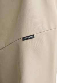 Beige stof met een gladde textuur, met een zichtbaar zwart label waarop "PETROL IND" staat, dicht bij de naad genaaid.