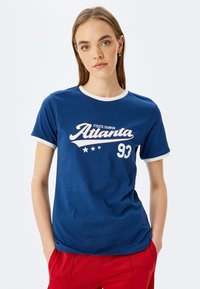 Sininen puuvillapaita, jossa on valkoinen reuna, ja jossa on tyylitelty teksti "Atlanta" ja "93". Pehmeä rakenne, rento malli.