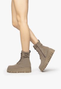 Bottines beiges avec une semelle plateforme épaisse, un laçage à l'avant, une fermeture éclair sur le côté et un matériau en daim texturé.