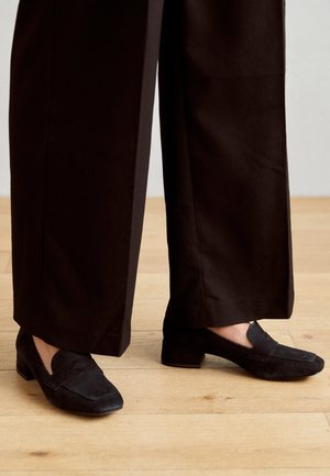 Czarne zamszowe loafersy z niskim obcasem, z przeszytym detalem w postaci kokardy na cholewce, zestawione z szerokimi czarnymi spodniami na drewnianej podłodze.