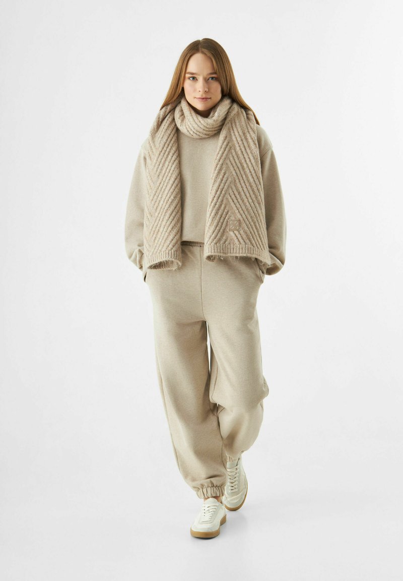 Jonge vrouw die naar voren loopt, gekleed in een beige gebreide sjaal, bijpassende sweatshirt, joggingbroek en witte sneakers, tegen een witte achtergrond.