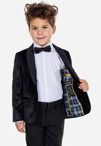 Blazer de terciopelo negro con solapas de satén, camisa blanca, pajarita negra y forro a cuadros. Los pantalones son oscuros, de corte ajustado.