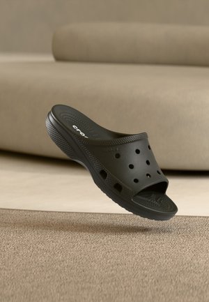 Fekete, könnyen felhúzható Crocs papucs szellőző lyukakkal, texturált talppal, bent szőnyegen, bézs kanapé mellett bemutatva.