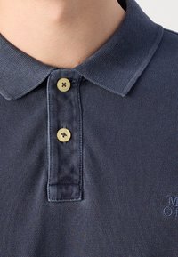 Gros plan sur une personne portant un polo bleu marine avec deux boutons beiges fixés et une partie de texte brodé visible.