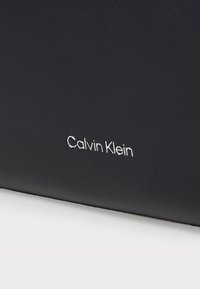 Portefeuille en cuir noir avec une surface texturée, présentant le logo embossé blanc "Calvin Klein" près du bas. Design minimaliste.