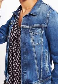 Jeansjacke in Hellblau mit klassischem Schnitt, ausgestattet mit Brusttasche, Knopfverschluss und ausgewaschenem Material; über einem gemusterten Oberteil getragen.