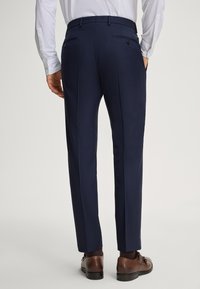 Pantaloni da donna color navy in tessuto intrecciato, vestibilità sartoriale, con doppie tasche posteriori con bottoni, abbinati a scarpe eleganti marroni e calzini scuri.
